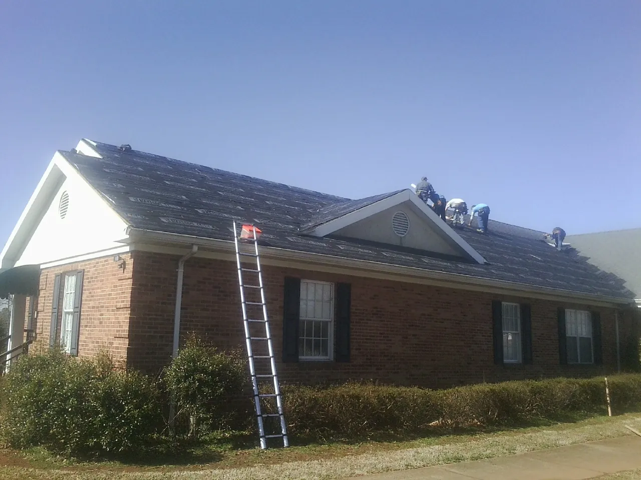 Crew tearing shingles off a brick ranch roof with a ladder set — Emergency Roof Repair work by our crew in Mattawan