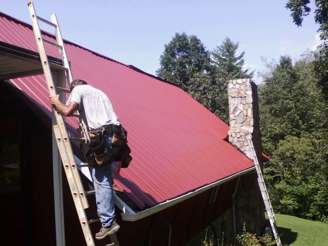 Roofer on a ladder beside a red metal roof and stone chimney — Soffit & Fascia Repair by Roofing Brothers in Mattawan